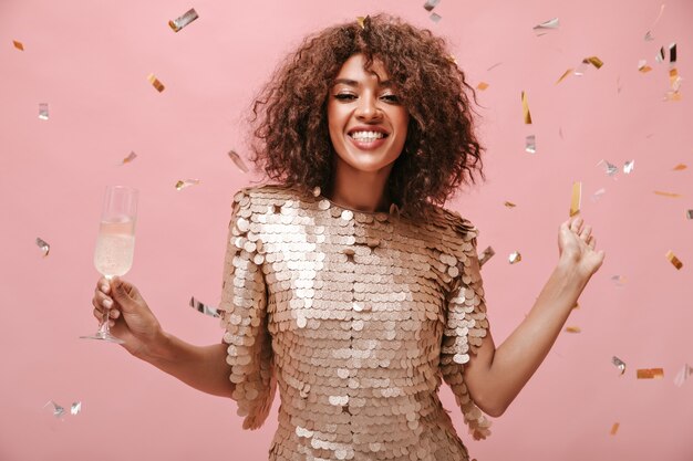 Femme brune positive en robe beige moderne souriante et posant avec un verre de vin et des confettis sur un mur rose.