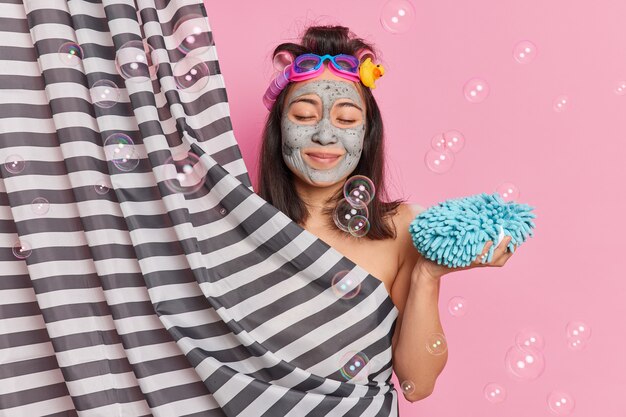 Une femme brune ravie garde les yeux fermés tient une éponge de bain prend une douche cache un corps nu derrière un rideau fait une coiffure frisée se soucie de la peau subit des procédures d'hygiène. Concept de douche