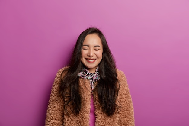 Femme Brune Ravie Avec Une Peau Saine, Sourit Sincèrement, Porte Un Manteau Brun, Garde Les Yeux Fermés, Isolée Sur Un Mur De Studio Lilas, S'amuse, Pose Pour La Photo. Gens