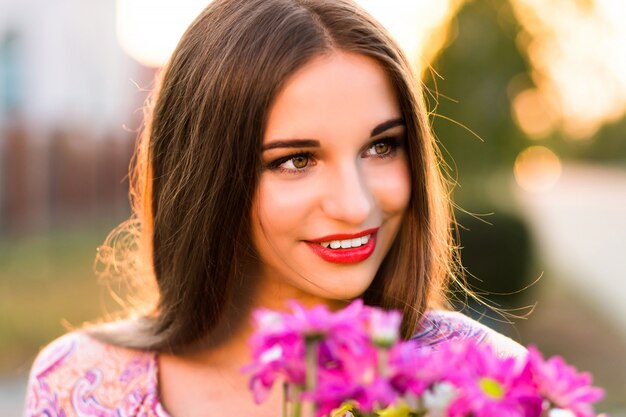 Femme brune sensuelle posant avec bouquet de fleurs après une date romantique, couleurs du coucher du soleil, robe élégante et maquillage.