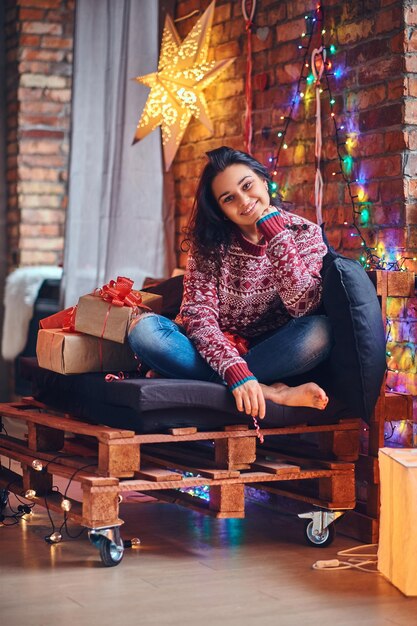 Femme brune sexy aux pieds nus vêtue d'un jean et d'un pull rouge posant sur un canapé dans une pièce avec une décoration de Noël.