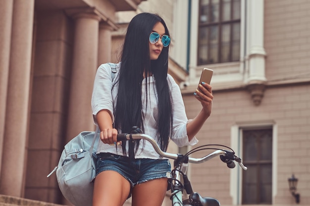 Une femme brune sexy portant un chemisier et un short en jean à lunettes de soleil, lisant le message sur un smartphone, debout sur les marches avec un vélo près d'un vieux bâtiment de la ville.
