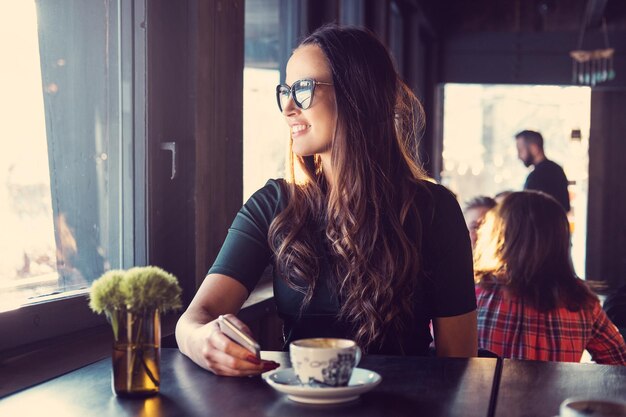 Femme brune souriante à l'aide d'un smartphone dans un café.