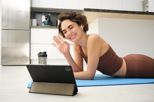 Femme brune souriante dans des vêtements de sport vagues main dit bonjour à la tablette enregistre vidéo blog sportif fitness
