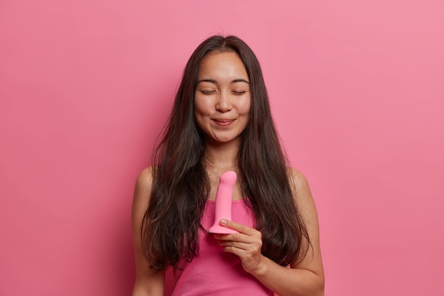 Une femme brune souriante heureuse pose avec un gode en silicone pour la pénétration sexuelle du vagin, de la bouche ou de l'anus, pas besoin de partenaire, stimule le clitoris, utilise des jouets sexuels pour la mastrubation, se tient à l'intérieur