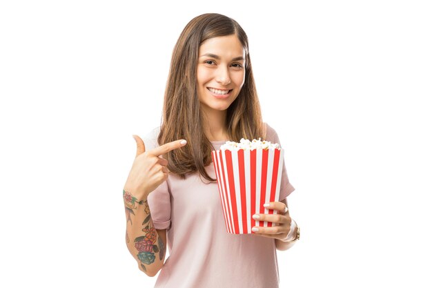 Femme brune souriante pointant sur un seau de pop-corn sur fond blanc