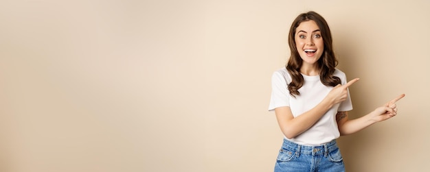 Femme brune souriante en t-shirt pointant les doigts vers la droite montrant une offre promotionnelle ou une publicité démontrant une bannière debout sur fond beige