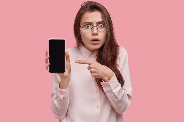 Femme brune stupéfaite tient un téléphone intelligent avec écran vide, pointe sur l'appareil, stupéfaite par d'énormes réductions, essaie de comprendre quelque chose