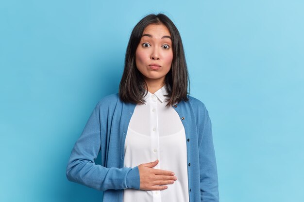 Une femme brune surprise ressent une gêne dans l'estomac après avoir mangé de la restauration rapide garde la main sur le ventre plein porte une chemise blanche et un pull décontracté