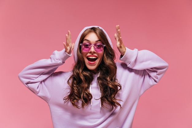 Une femme brune en sweat à capuche surdimensionné violet regarde la caméra sur un mur rose