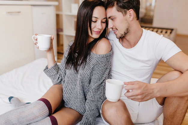 Femme brune timide en pull gris et demi-tuyau posant sur le lit avec son mari