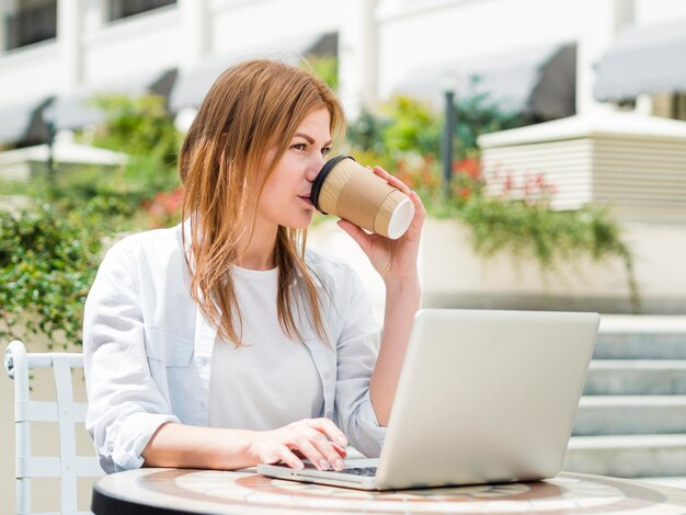 Femme buvant un café à l'extérieur tout en travaillant sur un ordinateur portable