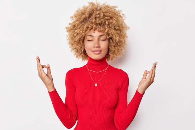 Photo gratuite une femme calme et concentrée aux cheveux bouclés garde les yeux fermés et médite à l'intérieur. le geste de mudra porte un col roulé rouge décontracté isolé sur fond blanc essaie de soulager le stress. pose zen