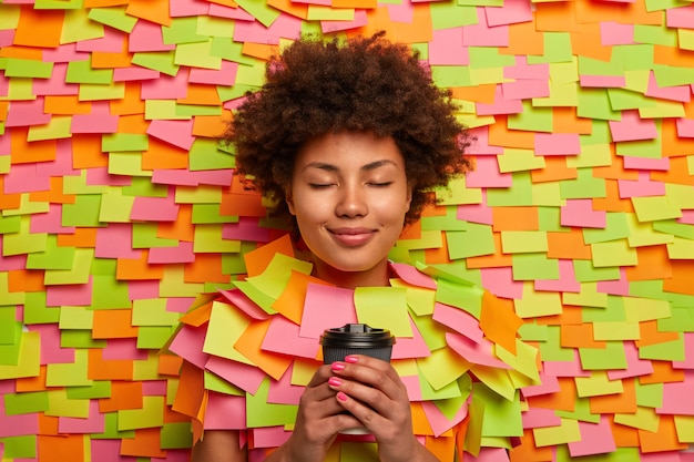 Photo gratuite une femme calme et détendue boit du café à emporter, garde les yeux fermés, pense à quelque chose d'agréable, aime boire une boisson rafraîchissante, entourée d'autocollants colorés. gens, concept de mode de vie