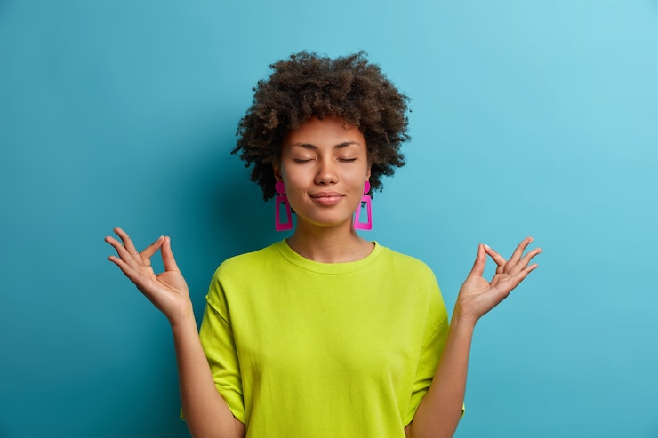Photo gratuit une femme calme et détendue à la peau sombre se tient en posture de lotus, se sent soulagée, essaie de se concentrer dans une atmosphère paisible, porte des vêtements verts décontractés, isolés sur un mur bleu. concept de langage corporel