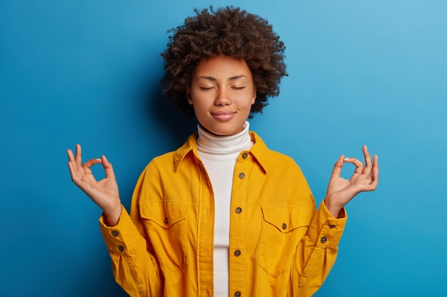 Photo gratuite femme calme à la peau sombre soulagée ferme les yeux, fait un geste mudra, vêtue d'une chemise jaune, se sent détendue, pose sur fond bleu