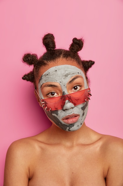 Une femme calme à la recherche sérieuse porte un masque de beauté, se tient torse nu à l'intérieur, porte des lunettes de soleil roses, réduit les rides et les points noirs, isolés sur un mur rose. Tir vertical. Rajeunissement, bien-être