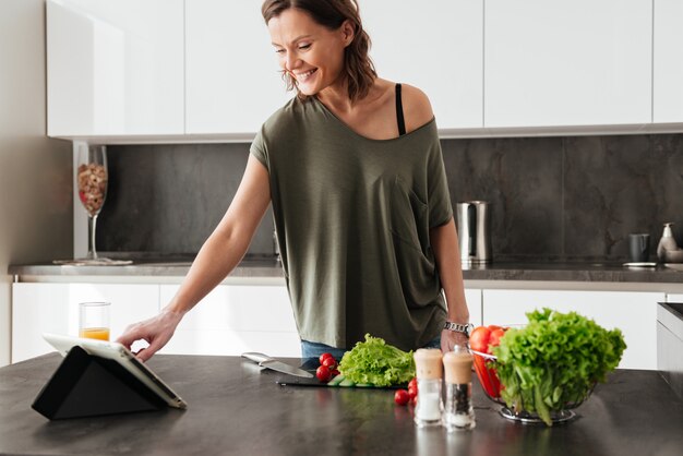 Femme casual souriante, debout près de la table avec des légumes dans la cuisine et à l'aide d'un ordinateur portable