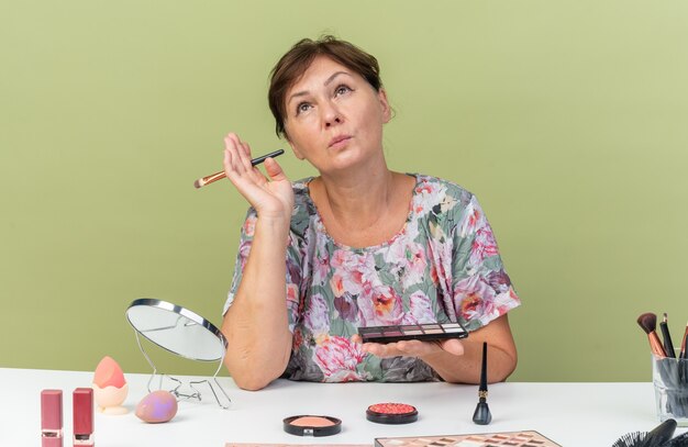 Femme caucasienne adulte confiante assise à table avec des outils de maquillage tenant une palette de fard à paupières et un pinceau de maquillage en levant