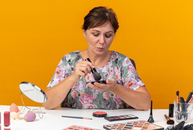 Une femme caucasienne adulte confiante assise à table avec des outils de maquillage tient un pinceau de maquillage et regarde le blush isolé sur un mur orange avec un espace de copie