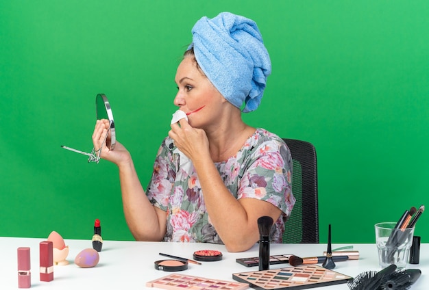 Une femme caucasienne adulte confiante aux cheveux enveloppés dans une serviette assise à table avec des outils de maquillage s'essuie la bouche avec une serviette humide isolée sur un mur vert avec espace de copie