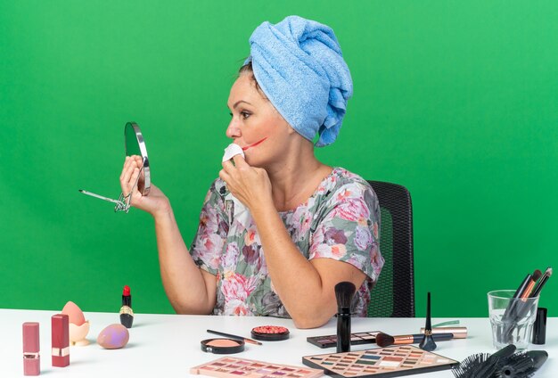 Une femme caucasienne adulte confiante aux cheveux enveloppés dans une serviette assise à table avec des outils de maquillage s'essuie la bouche avec une serviette humide isolée sur un mur vert avec espace de copie