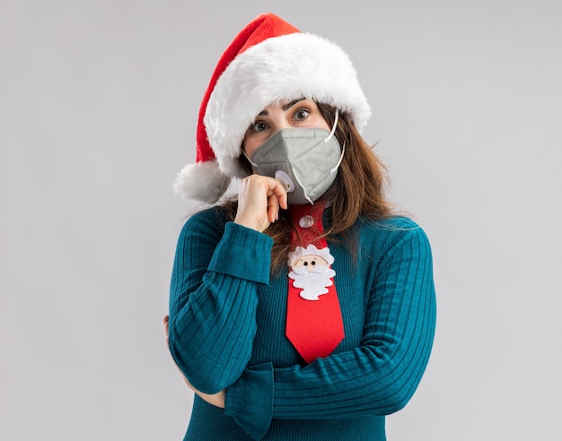 Femme caucasienne adulte inquiète avec bonnet de Noel et cravate de Noel portant un masque médical isolé sur mur blanc avec espace de copie