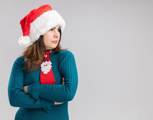 Femme caucasienne adulte sans idée avec bonnet de noel et cravate de noel se tient avec les bras croisés regardant de côté isolé sur mur blanc avec espace de copie