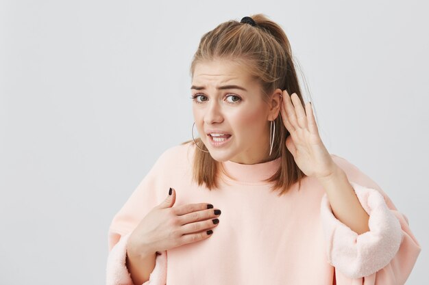 Femme caucasienne agressive en colère en pull rose regardant avec colère et appel, se montrant, tenant la main derrière son oreille comme si elle n'entend pas ce qu'on lui dit de faire, férocement à écouter.