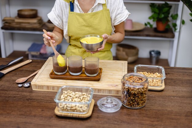 Femme caucasienne dans la cuisine fait des puddings de chia avec de la confiture de mangue. Désert fait de lait d'amande, graines de chia, cacao, confiture de mangue et granola.