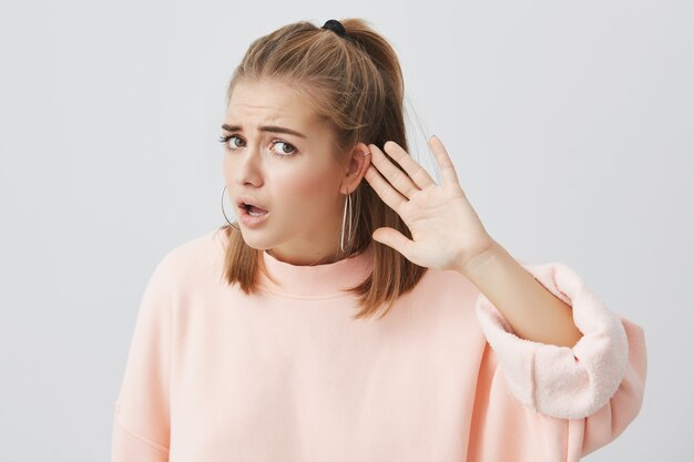 Femme caucasienne désemparée en pull rose à la recherche avec une expression confuse et perplexe, fronçant les sourcils, tenant la main derrière l'oreille comme si elle n'entend pas ce qu'on lui dit de faire, faisant semblant de le faire