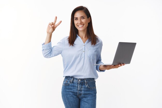 Femme caucasienne gaie et insouciante professionnelle des années 30 avec tatouage travaillant, se sentir confiante, le projet se passe bien, montrer le signe de paix de la victoire, tenir un ordinateur portable, tenir le mur blanc satisfait, assurer la finition à temps