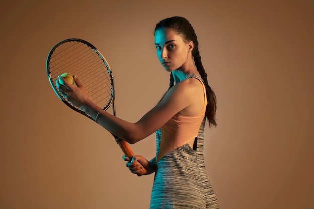 Une femme caucasienne jouant au tennis isolé sur fond marron en lumière mixte et néon. Ajuster la jeune joueuse en mouvement ou en action pendant le jeu de sport. Concept de mouvement, sport, mode de vie sain.