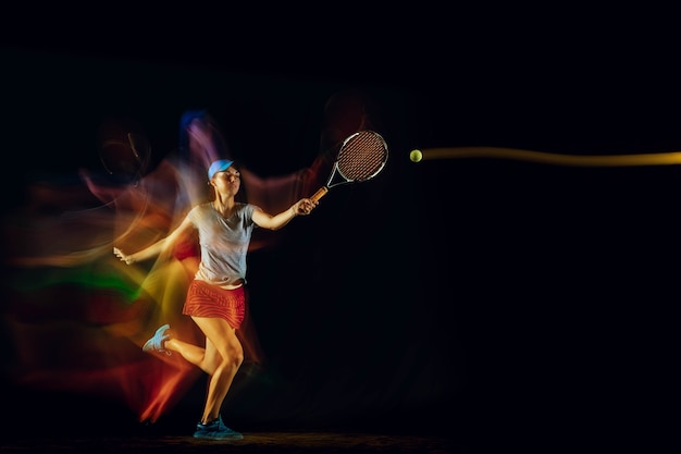 Photo gratuite une femme caucasienne jouant au tennis isolé sur un mur noir en lumière mixte et stobe. ajuster la jeune joueuse en mouvement ou en action pendant le jeu de sport. concept de mouvement, sport, mode de vie sain.