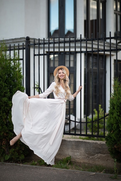 Une femme caucasienne joyeuse aux longs cheveux blonds, aux yeux bleus et au sourire parfait en longue robe blanche avec un chapeau se promène à l'extérieur près des maisons