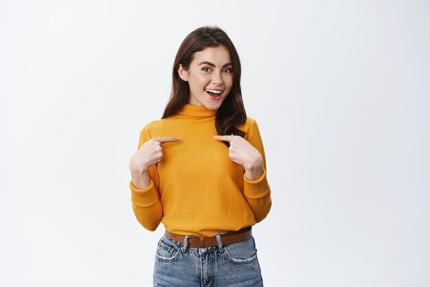 Femme caucasienne souriante pointant vers elle-même et s'autopromouvant introduire ou louer sa propre réalisation debout sur fond blanc confiant