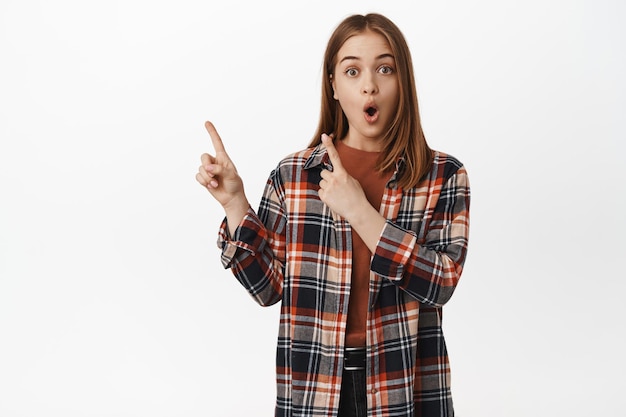 Une femme caucasienne surprise haletant d'admiration, pointant vers le coin supérieur gauche avec une expression de visage impressionnée, montrant un texte promotionnel de réduction, une publicité, un fond blanc