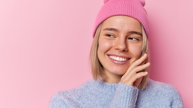 Une femme caucasienne tendre et heureuse touche le visage, sourit doucement, regarde à pleines dents et admire quelque chose qui a une peau propre et saine, porte un chapeau et un pull isolé sur fond rose, espace vide