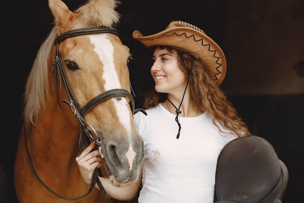 Femme de cavalier tenant une selle dans une écurie. La femme a les cheveux longs et un t-shirt blanc