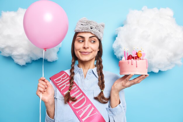 une femme célèbre son 26e anniversaire dans un cercle familial tient un ballon d'hélium gonflé pour un gâteau de fête porte une chemise avec un ruban se prépare pour une fête isolée sur bleu