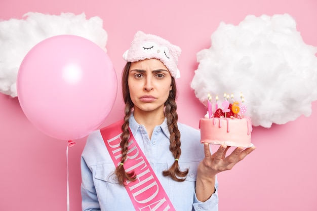 une femme célèbre son 26e anniversaire seule détient un gâteau aux fraises et un ballon gonflé vêtu de vêtements domestiques décontractés isolés sur rose