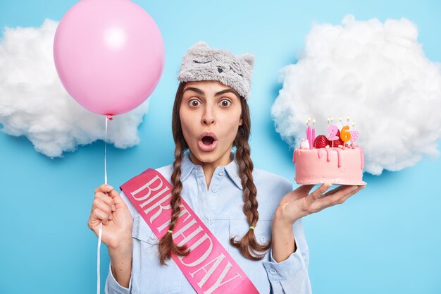 une femme célèbre son anniversaire pose avec un gâteau et un ballon gonflé garde la bouche ouverte vêtue d'une chemise et d'un masque de sommeil sur la tête pose contre le bleu