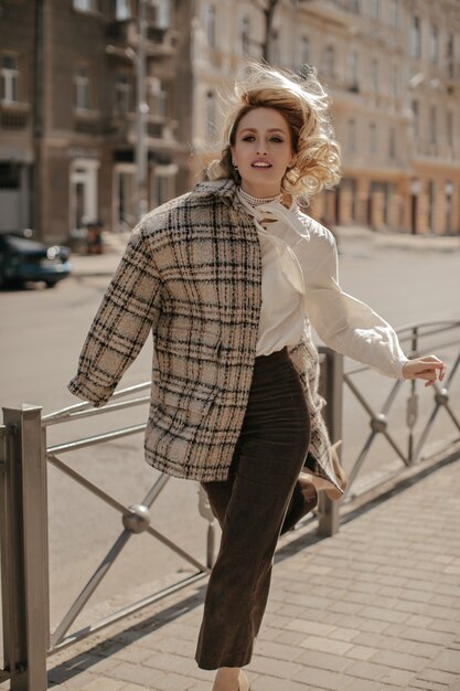 Femme charmante blonde bouclée dans un pantalon marron élégant, un chemisier blanc et un manteau à carreaux courant dans le centre-ville