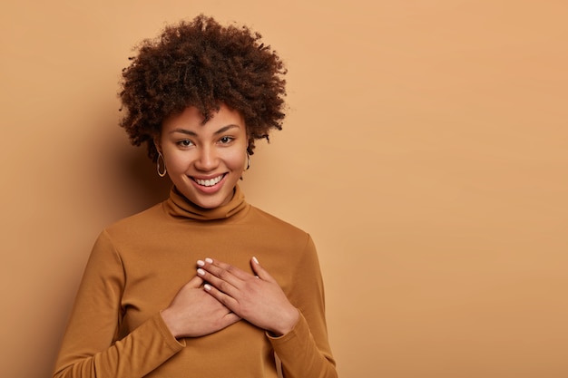 Photo gratuite une femme charmante et heureuse avec une coiffure afro reçoit un beau cadeau, serre les mains contre le cœur, apprécie l'aide, sourit doucement, se sent très reconnaissante