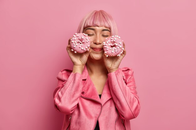 Femme charmante satisfaite aux cheveux roses et à la frange, ferme les yeux, imagine un goût agréable de beignets, vêtue d'une veste rose