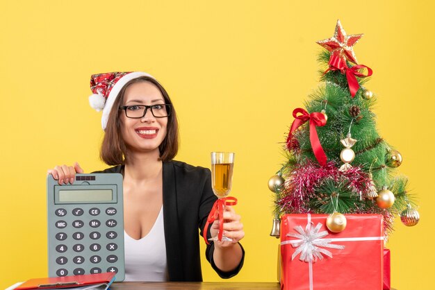 Femme charmante souriante en costume avec chapeau de père Noël et lunettes montrant calculatrice et élevage de vin au bureau sur jaune isolé
