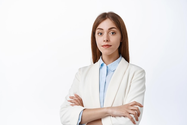 Une femme chef de la direction confiante en costume d'affaires croise les bras sur la poitrine, elle a l'air déterminée et prête à la caméra, debout comme un fond blanc professionnel