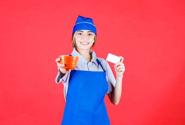 Femme chef en tablier bleu tenant une tasse de nouilles en céramique orange et présentant sa carte de visite