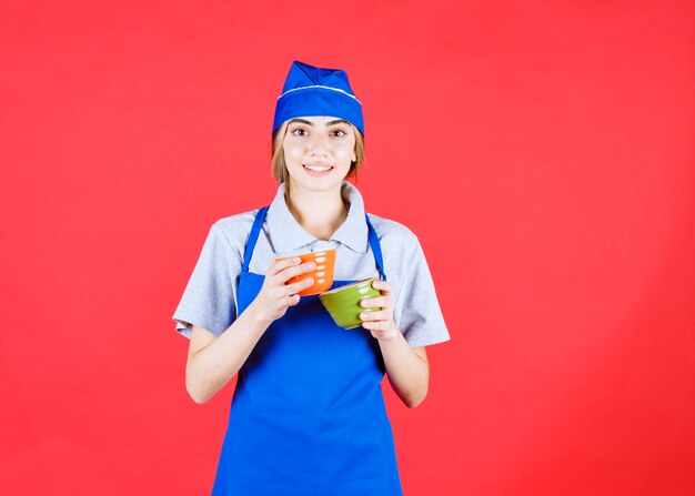 Femme chef en tablier bleu tenant des tasses de nouilles en céramique orange et vert