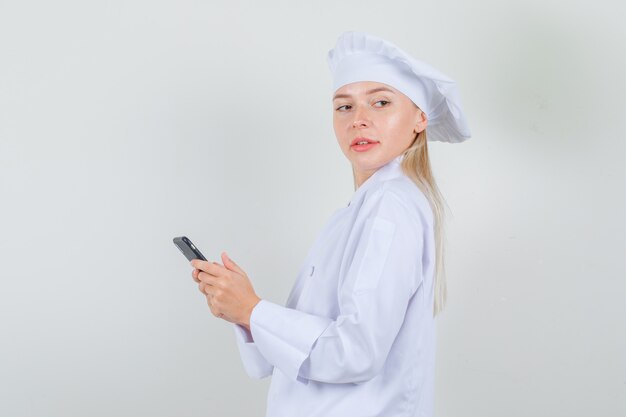 Femme chef tenant le smartphone et regardant de côté en uniforme blanc